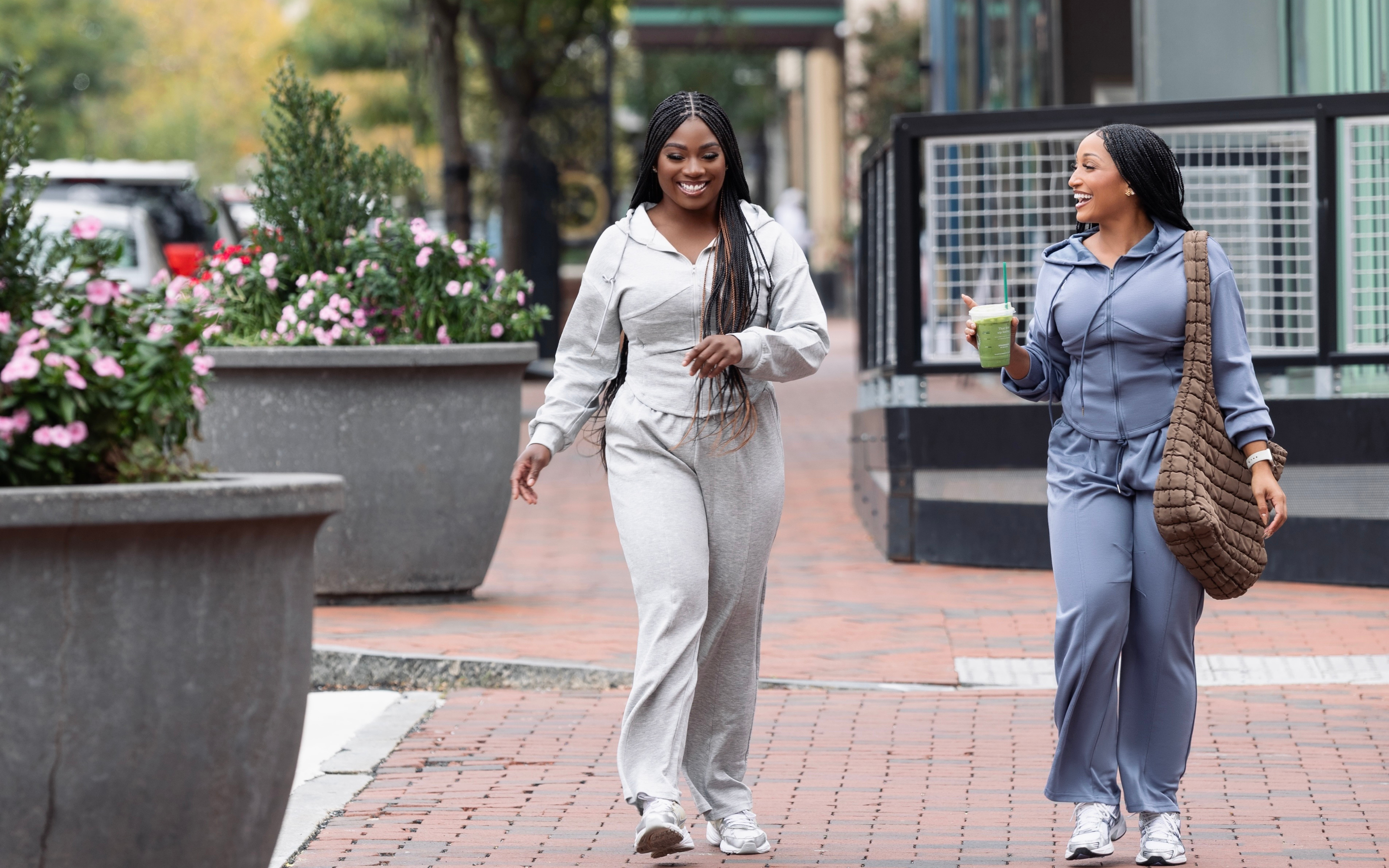 Two women walking outdoors on a sidewalk with plants and a building in the background.